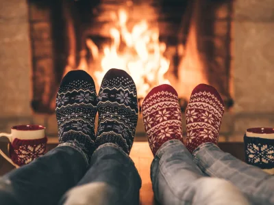 Two people in festive socks sitting by a cozy fire with mugs of hot drinks.