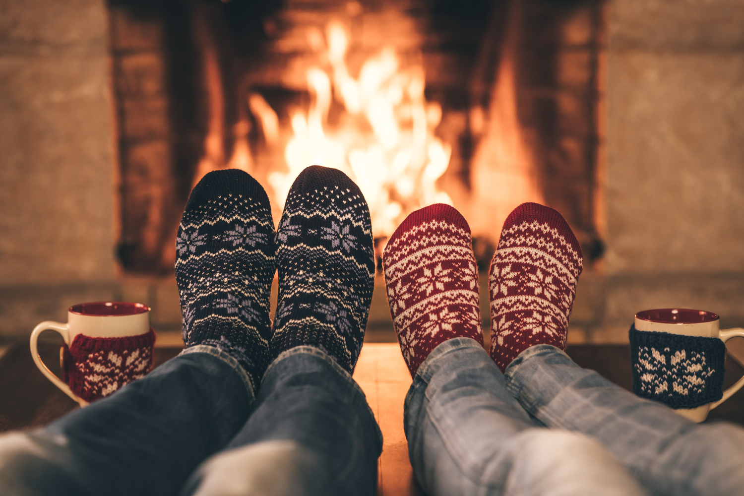 Two people in festive socks sitting by a cozy fire with mugs of hot drinks.